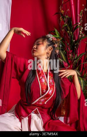 Femme asiatique en robe rouge traditionnelle portant la couronne de fleur assise avec les yeux fermés et le bras levé à côté de la plante d'intérieur dans le studio Banque D'Images