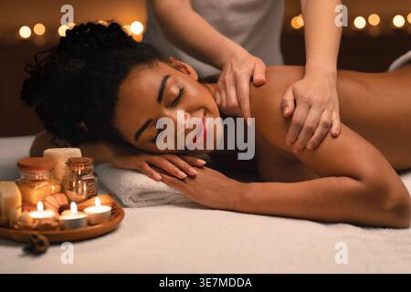 Femme noire avec les yeux fermés ayant massage de l'épaule couchée sur le lit près de bougies allumées dans le spa Banque D'Images