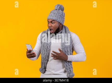 Homme noir choqué portant un bonnet tricoté et une écharpe regardant un smartphone debout sur fond jaune en studio Banque D'Images