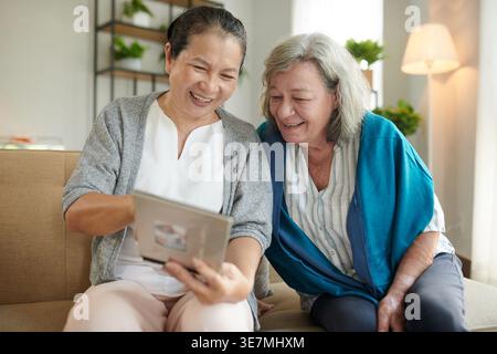 Femme asiatique d'âge moyen montrant un album photo à une amie caucasienne senior assise dans le salon pendant la journée Banque D'Images