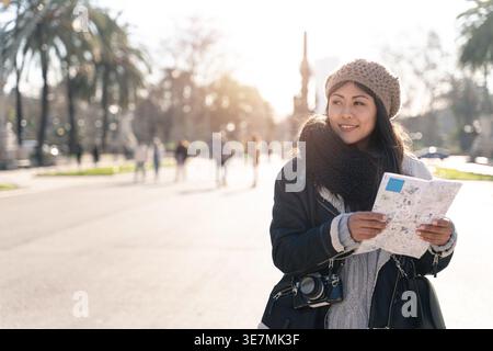 Souriant touriste asiatique dans des vêtements chauds tenant la carte de la ville et regardant loin tout en se tenant debout avec appareil photo et sac sur la rue de la ville pendant vaca d'hiver... Banque D'Images