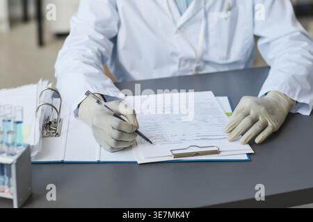 Gros plan d'une technologue médicale noire portant une blouse de laboratoire écrivant des notes assis à table dans un laboratoire médical pendant la journée Banque D'Images