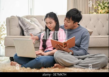 Homme asiatique tenant le livre aidant la petite amie asiatique travaillant sur ordinateur portable assis sur le tapis de sol avec les jambes croisées appuyées contre le canapé dans le salon à ... Banque D'Images