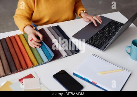 Mains de concepteur féminin mixte travaillant sur ordinateur portable choisissant la couleur pour les meubles tout en étant assis à table dans le bureau Banque D'Images