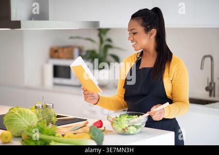 Femme noire souriante portant tablier lisant la recette sur le cahier faisant de la salade tout en se tenant debout à table dans la cuisine à la maison Banque D'Images