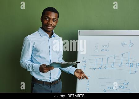 Enseignant noir tenant le presse-papiers et expliquant les notes de musique écrites sur le tableau blanc tout en se tenant debout sur fond vert dans le studio Banque D'Images