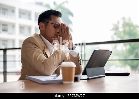 Homme d'affaires asiatique frustré avec les mains priantes assis avec les yeux fermés à la table en bois avec des documents, tablette, et café sur le balcon dans son... Banque D'Images