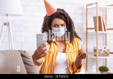 Bouleversé femme noire portant chapeau de fête et masque médical tenant cupcake avec bougie et ayant appel vidéo sur smartphone tout en étant assis sur le canapé dans Livin... Banque D'Images