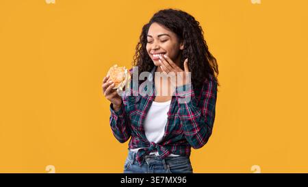 Femme noire aux cheveux bouclés mangeant un hamburger riant et essuyant les lèvres tout en se tenant debout sur fond jaune en studio Banque D'Images