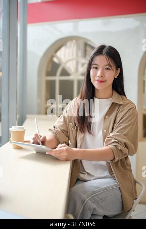 Étudiante asiatique tenant un stylet et une tablette assis à table près de la fenêtre dans le café Banque D'Images