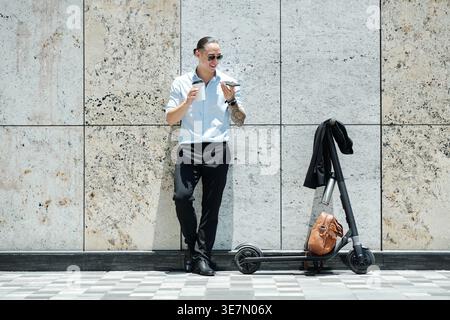 Homme d'affaires mixte portant des lunettes de soleil tenant une tasse de café et parlant au smartphone tout en se tenant à côté du scooter électrique sur la rue de la ville Banque D'Images