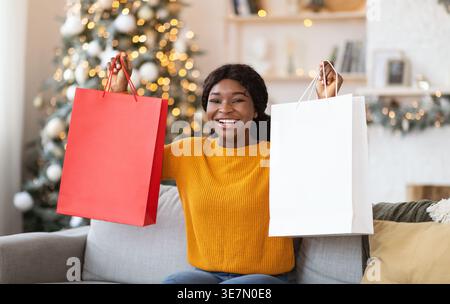 Joyeuse femme noire tenant des sacs à provisions tout en étant assise sur le canapé dans le salon à la maison Banque D'Images