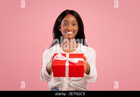 Femme noire souriante avec de longs cheveux droits tenant la boîte cadeau rouge tout en se tenant debout sur fond rose dans le studio Banque D'Images