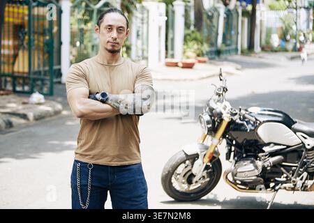 Homme de race mixte avec les bras croisés debout près de la moto sur la rue le jour ensoleillé Banque D'Images