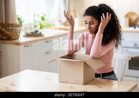 Bouleversée femme du moyen-Orient faisant des gestes des mains regardant la boîte en carton ayant un problème avec son paquet tout en étant assis à table dans la cuisine à la maison Banque D'Images
