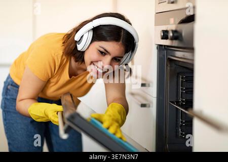 Adolescente hispanique portant des écouteurs et des gants en caoutchouc en utilisant un chiffon dans le four de nettoyage tout en se tenant debout dans la cuisine à la maison Banque D'Images