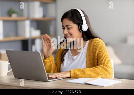 Femme du moyen-Orient souriante portant des écouteurs agitant la main et ayant un appel vidéo sur ordinateur portable tout en étant assis au bureau avec ordinateur portable et tasse de café je... Banque D'Images