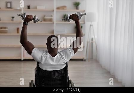 Vue arrière de l'homme chauve noir handicapé en fauteuil roulant s'exerçant avec des haltères dans le salon à la maison Banque D'Images