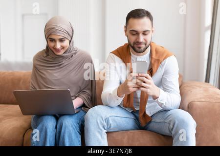 Femme musulmane arabe en hijab et mari caucasien utilisant un ordinateur portable et un smartphone tout en étant assis ensemble sur un canapé dans le salon à la maison Banque D'Images