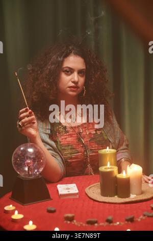 Diseuse de bonne fortune féminine de race mixte tenant un bâton brûlant tout en étant assise à table avec boule de cristal et cartes de tarot à l'intérieur Banque D'Images
