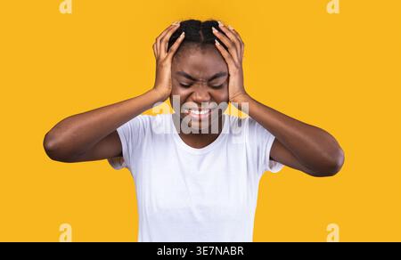 Femme noire tenant sa tête souffrant de douleur tout en se tenant debout avec les yeux fermés sur fond jaune dans le studio Banque D'Images