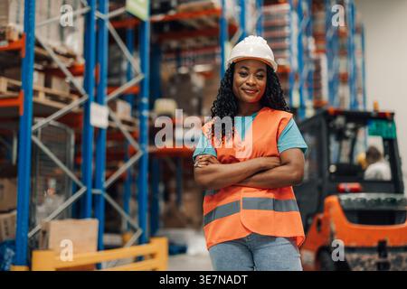 Travailleuse noire avec les bras croisés portant un casque de sécurité et un gilet de sécurité debout dans l'entrepôt pendant le travail Banque D'Images