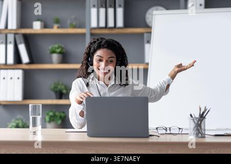 Enseignante noire portant des écouteurs avec les bras levés ayant cours en ligne assis à table au bureau à domicile Banque D'Images