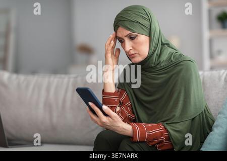 Frustrée femme musulmane d'âge moyen-oriental en hijab touchant sa tête et utilisant un smartphone tout en étant assise sur un canapé dans le salon à la maison Banque D'Images