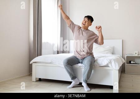 Homme asiatique étirant ses bras après s'être réveillé tout en étant assis sur le lit dans la chambre à coucher à la maison Banque D'Images