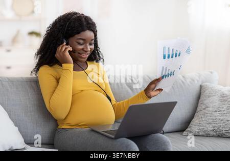 Femme enceinte noire avec des cheveux bouclés portant chemise jaune à manches longues travaillant à l'aide d'un casque ayant videocall sur ordinateur portable assis sur le canapé dans la... Banque D'Images