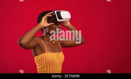 Femme noire joyeuse avec les cheveux courts portant un casque de réalité virtuelle tout en se tenant devant un fond rouge en studio Banque D'Images