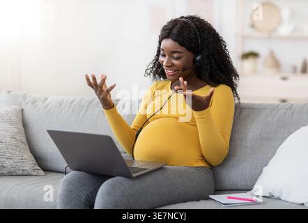 Femme enceinte noire avec des cheveux bouclés portant chemise à manches longues jaune gestuelle des mains tout en travaillant à l'aide d'un casque ayant videocall sur ordinateur portable sittin... Banque D'Images