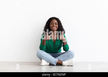 Femme noire souriante montrant les pouces assis avec les jambes croisées sur le sol sur fond blanc dans le studio Banque D'Images