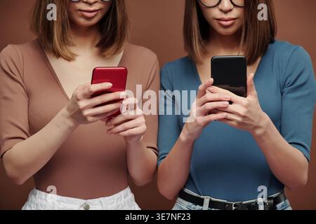 Sœurs asiatiques portant des lunettes à l'aide de smartphones debout sur fond brun en studio Banque D'Images