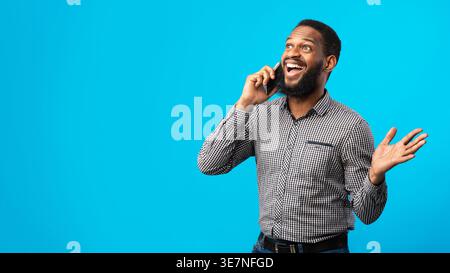 Homme noir surpris ayant un appel téléphonique regardant vers le haut tout en se tenant debout sur fond bleu dans le studio Banque D'Images