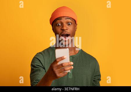 Homme noir portant un bonnet avec une expression choquée à l'aide d'un smartphone tout en se tenant sur fond jaune dans le studio Banque D'Images