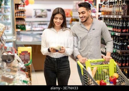 Couple moyen-oriental vérifiant les légumes tout en se tenant debout avec chariot dans le supermarché Banque D'Images