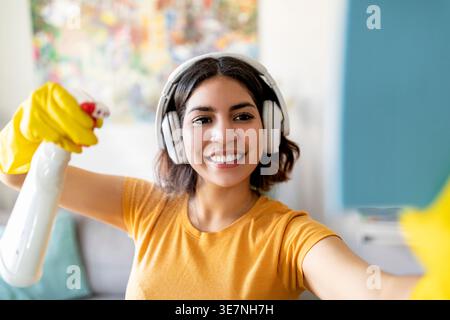 Adolescente hispanique portant des écouteurs et des gants en caoutchouc en utilisant un pulvérisateur et un chiffon dans les fournitures de nettoyage à la maison Banque D'Images