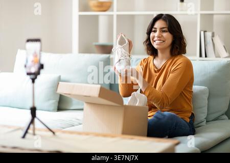 Hispanique femme vlogger enregistrant la vidéo tout en déballant le colis avec des chaussures de sneaker assis sur le lit dans la chambre à coucher à la maison Banque D'Images
