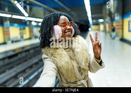 Femme noire joyeuse avec les cheveux bouclés portant des cache-oreilles et des lunettes de soleil prenant selfie montrant le signe de la paix tout en se tenant debout à la gare Banque D'Images