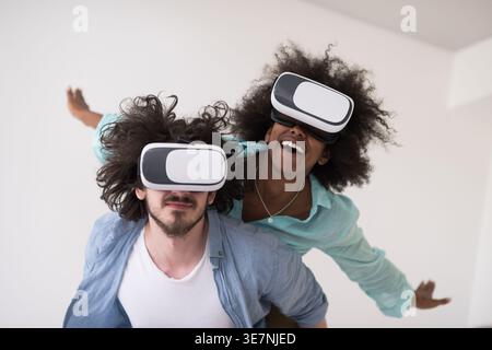 Couple noir et caucasien portant des casques VR ayant un plaisir piggyback équitation et expérimentant la réalité virtuelle sur fond blanc en studio Banque D'Images