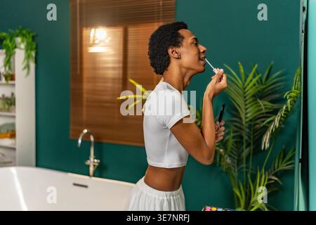 Homme gay hispanique regardant un miroir et mettant du rouge à lèvres tout en se tenant dans la salle de bain à la maison Banque D'Images