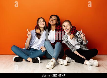 Adolescente noire embrassant des amis féminins caucasiens assis sur le sol sur fond orange dans le studio Banque D'Images
