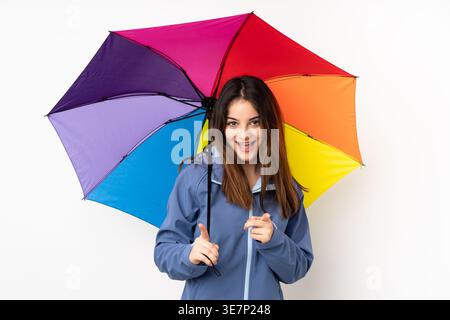 Femme hispanique portant une veste tenant un parapluie arc-en-ciel et pointant le doigt vers l'avant tout en se tenant debout sur fond blanc dans le studio Banque D'Images