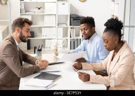 Couple noir sérieux lisant des documents ayant une réunion avec un agent masculin caucasien assis en face de la table dans le bureau Banque D'Images