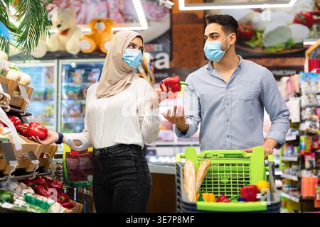 Couple souriant du moyen-Orient portant un masque facial choisissant des légumes sur des étagères tout en se tenant debout avec un chariot dans un supermarché Banque D'Images