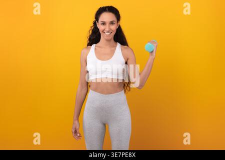 Femme du moyen-Orient souriante avec des cheveux bouclés portant des vêtements de sport tenant un haltère debout sur fond jaune dans le studio Banque D'Images