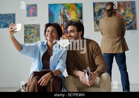 Couple noir et moyen-oriental prenant selfie à l'aide d'un smartphone tout en étant assis sur le banc dans la galerie d'art. Banque D'Images