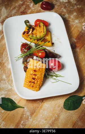 Plateau de légumes grillés gastronomiques avec poivrons de courgettes de maïs sucrées et tomates cerises servis sur une assiette blanche sur un bois rustique Banque D'Images