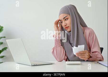 Femme asiatique en foulard calculant les dépenses à partir des factures regardant l'ordinateur portable tout en étant assis à table dans le bureau Banque D'Images